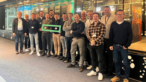 16 mannen staan als groep bij elkaar en kijken lachend de camera in. Ze houden een logo vast van Hewlett Packard Enterprise. Ze zijn onderdeel van de Wentzo IoT-trip naar Genève.