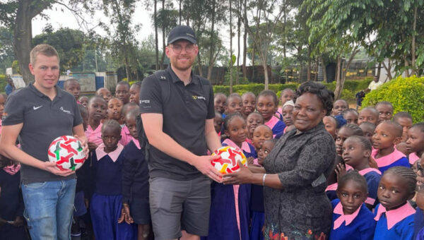 Foto van de Wentzo goes Kenya reis 2024. Twee Wentzo collega's houden een voetbal vast. Een overhandigt deze aan een lerares. Om hun heen staan kinderen van de basisschool.