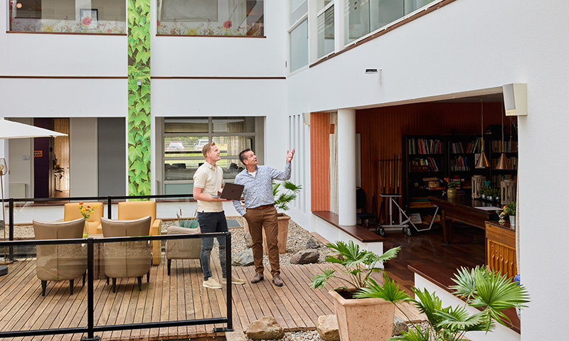 Twee personen kijken naar een netwerkapparatuur aan de muur in een lichte, moderne ruimte met houten vloer en planten.